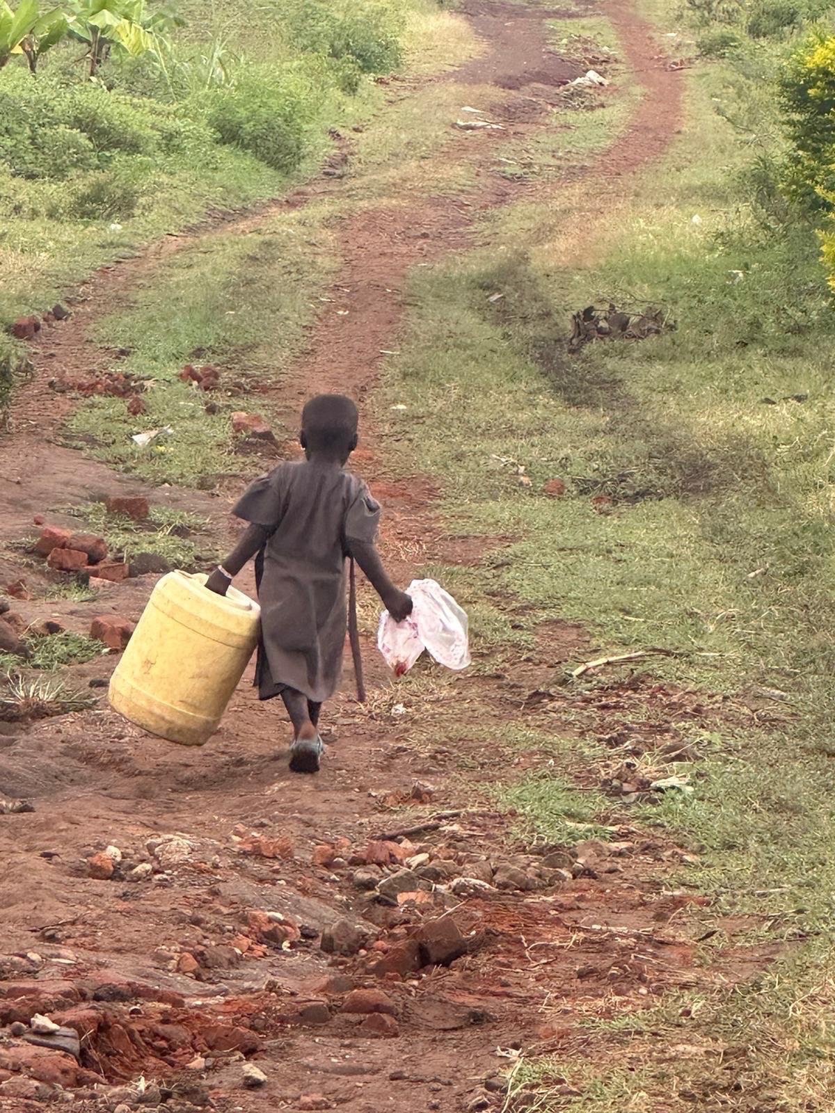 Child carrying a large yellow water container along a rural red-earth path in Uganda.