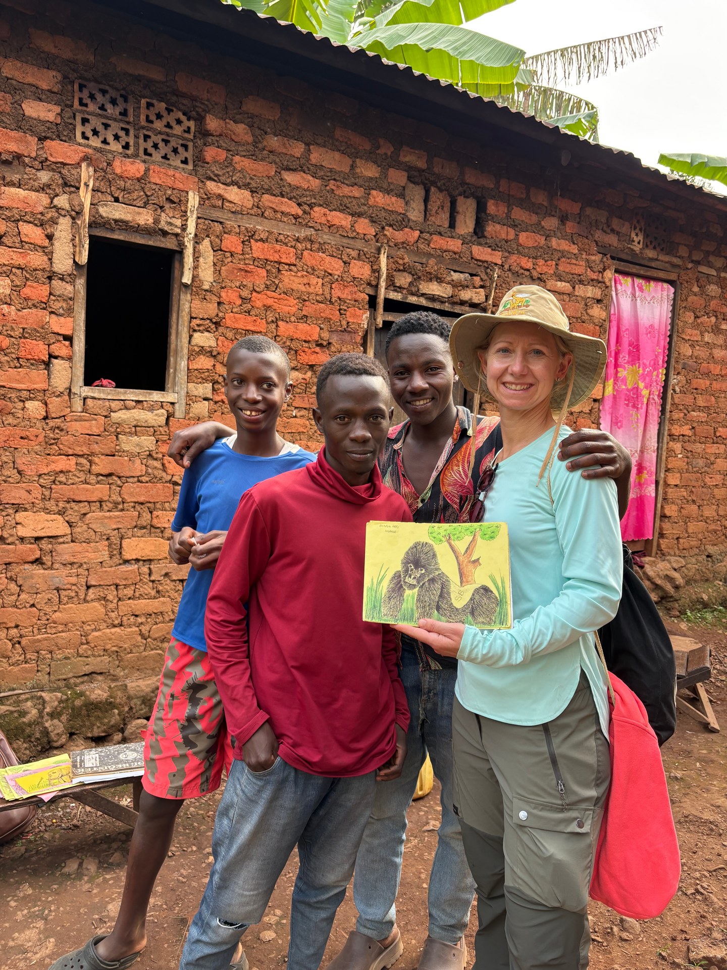 Clinton, a 17-year-old artist from Buhoma, stands with his family outside Gorilla Bluff Lodge selling hand-painted gorilla pictures. Photo: Mark Suer, January 2026. GPS: -0.9791°N, 29.6180°E.
