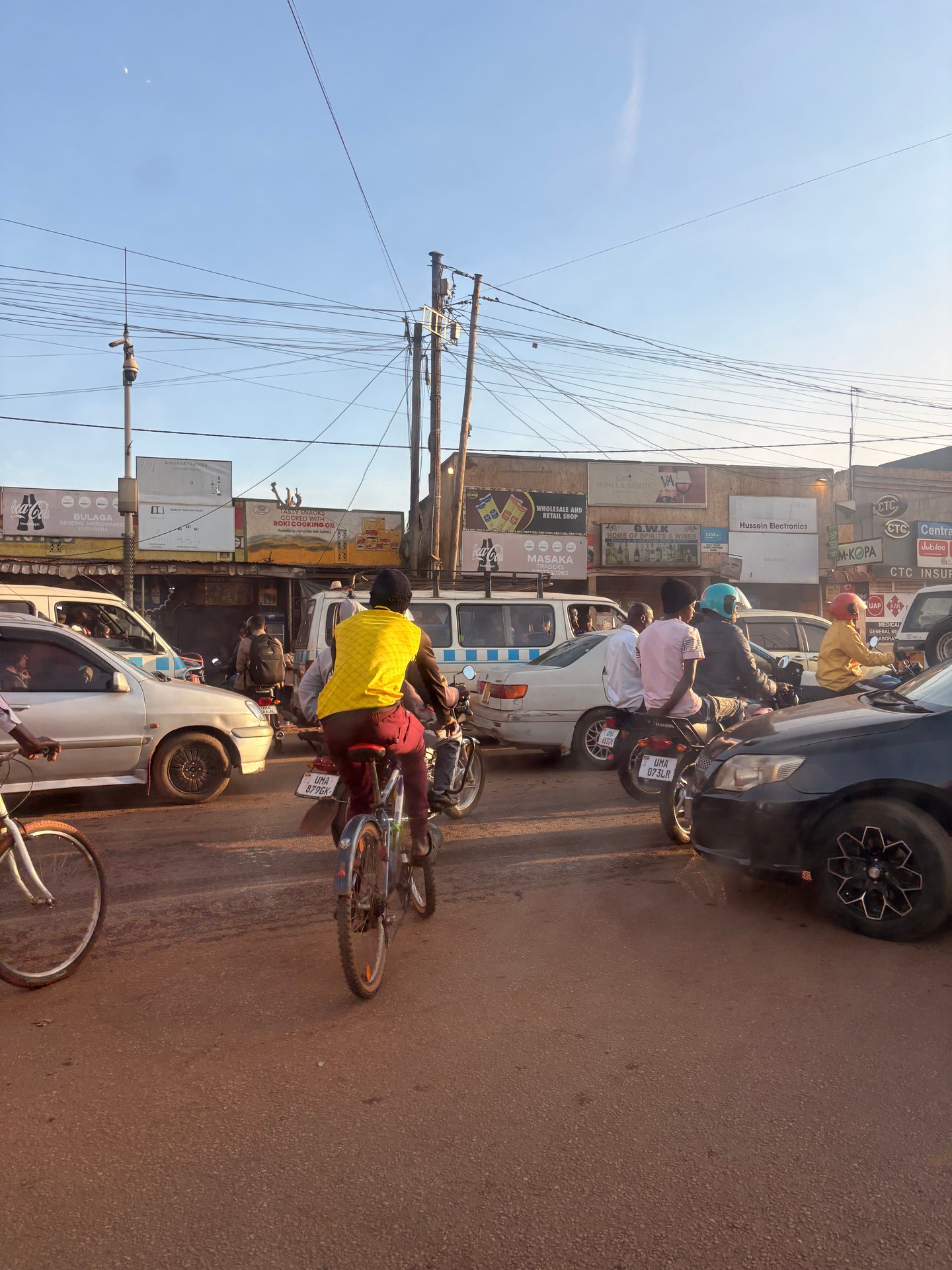 Bodaboda motorcycles, bicycles, and cars in Kampala city traffic, Uganda. Photo: Mark Suer, May 2026. GPS: 0.2917°N, 32.4996°E.