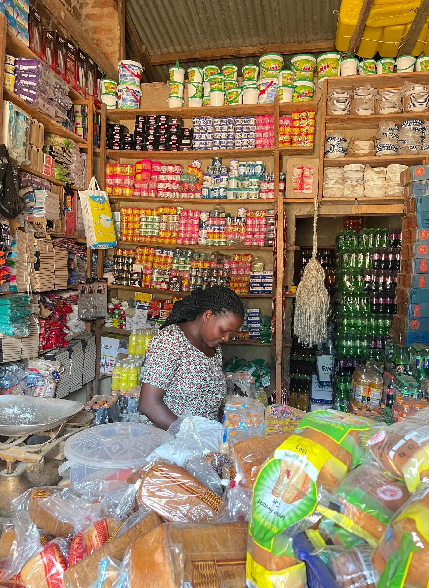A woman shopkeeper behind the counter of her small general store in Buhoma, shelves stocked with everyday goods, January 2026