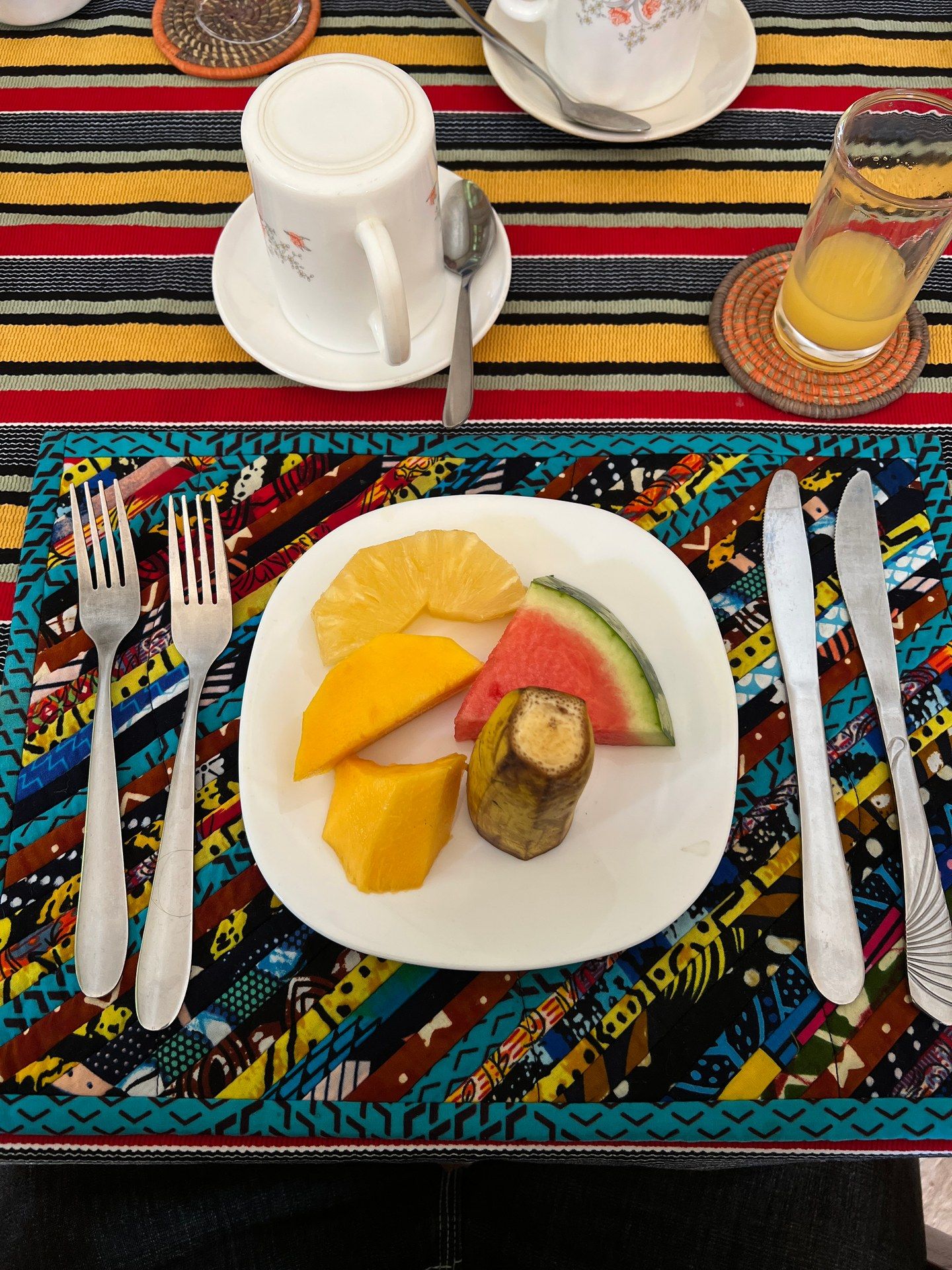 Simple breakfast at Gorilla Bluff Lodge in Buhoma: fresh mango, banana and melon on a white plate with a jug of fruit juice and cutlery, January 2026. Photo: Mark Suer.
