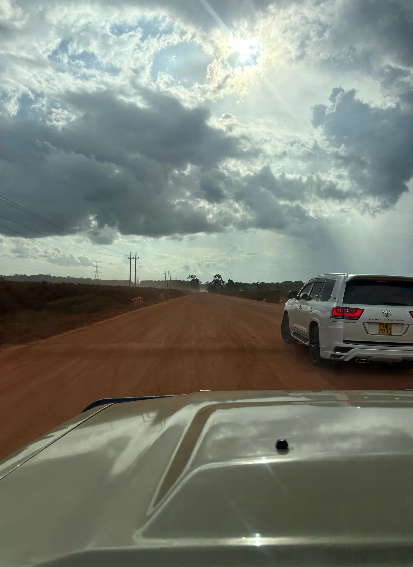 A white safari jeep travels the dusty Masaka Highway through Butambala, Uganda, with dramatic sunlight breaking through clouds. Photo: Mark Suer, January 2026