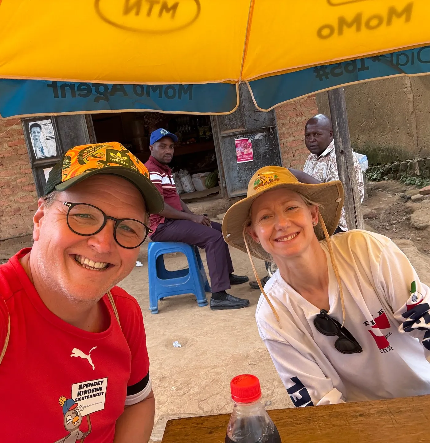 Two travellers rest on a bench outside a small shop on Buhoma's main road, drinking water in the morning, June 2026. Photo: Mark Suer