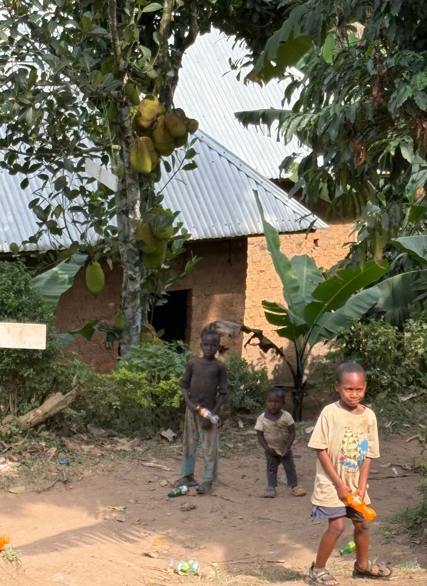 Three children from the neighborhood near the Buhoma orphanage stand before a simple building with a corrugated iron roof. Buhoma village, June 2026.